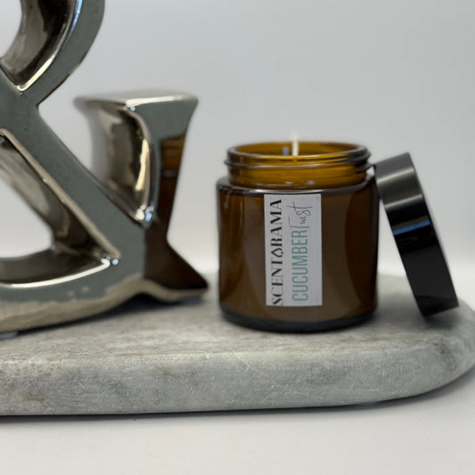 Amber candle jar in Cucumber Twist scent with a Scentarama label on a gray surface with a decorative metal ampersand symbol.