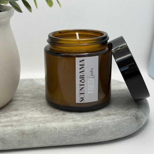 Amber candle jar in Fresh Linen Scent with black lid on a marble surface next to a plant