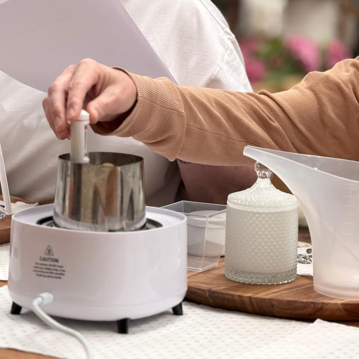 A person in a tan long-sleeve shirt operates a white wax melting pot with a metal thermometer, while another person in a white shirt looks on and holds papers. A glass jar of white wax and a translucent pouring cup sit on a wooden board on the table.