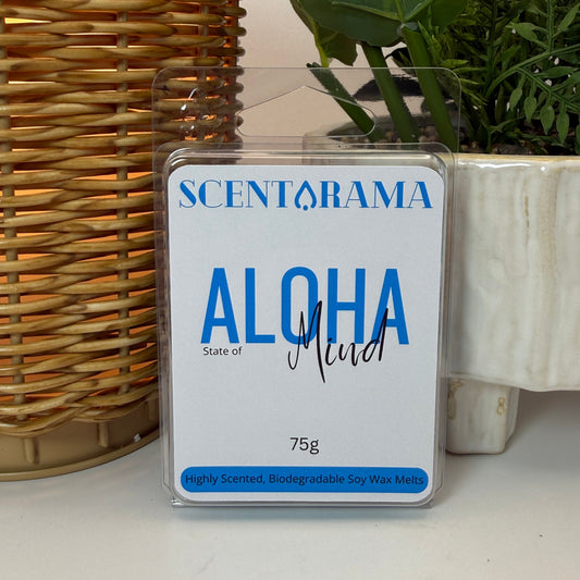 A 75g pack of Scentarama 'Aloha State of Mind' soy wax melts in a clear plastic clamshell package, displayed on a white surface next to a wicker basket and a potted plant.