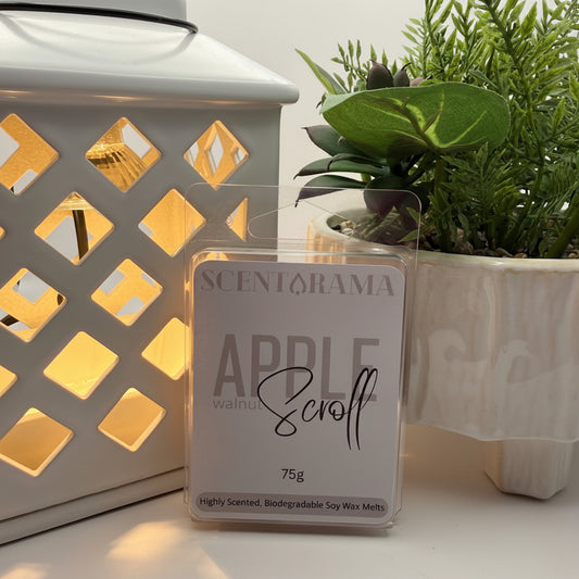 A 75g package of Scentarama Apple and walnut Scroll soy wax melts stands on a white surface, styled with a lit white lantern wax melt warmer on the left and a green potted plant on the right.