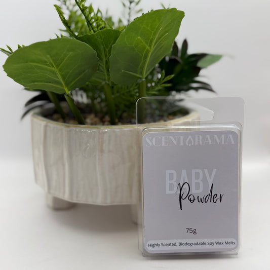A package of Scentarama Baby Powder 75g soy wax melts stands in front of a potted green plant on a white surface against a white background.