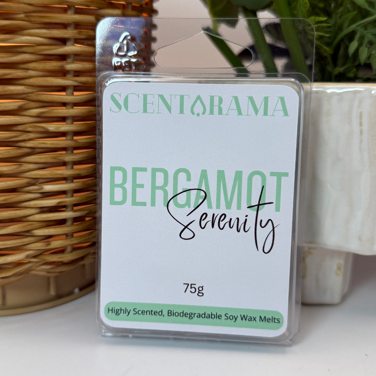 A packaged wax melt product labeled SCENTARAMA BERGAMOT Serenity, 75g, standing upright on a white surface with a wicker wax warmer and potted plants in the background.