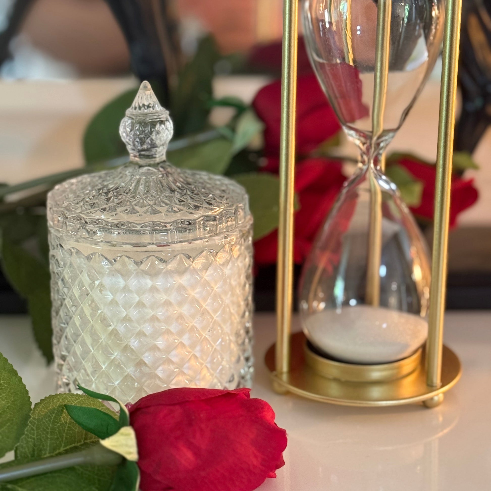 Decorative glass jar with a lid and gold hourglass on a surface with red roses and green leaves.