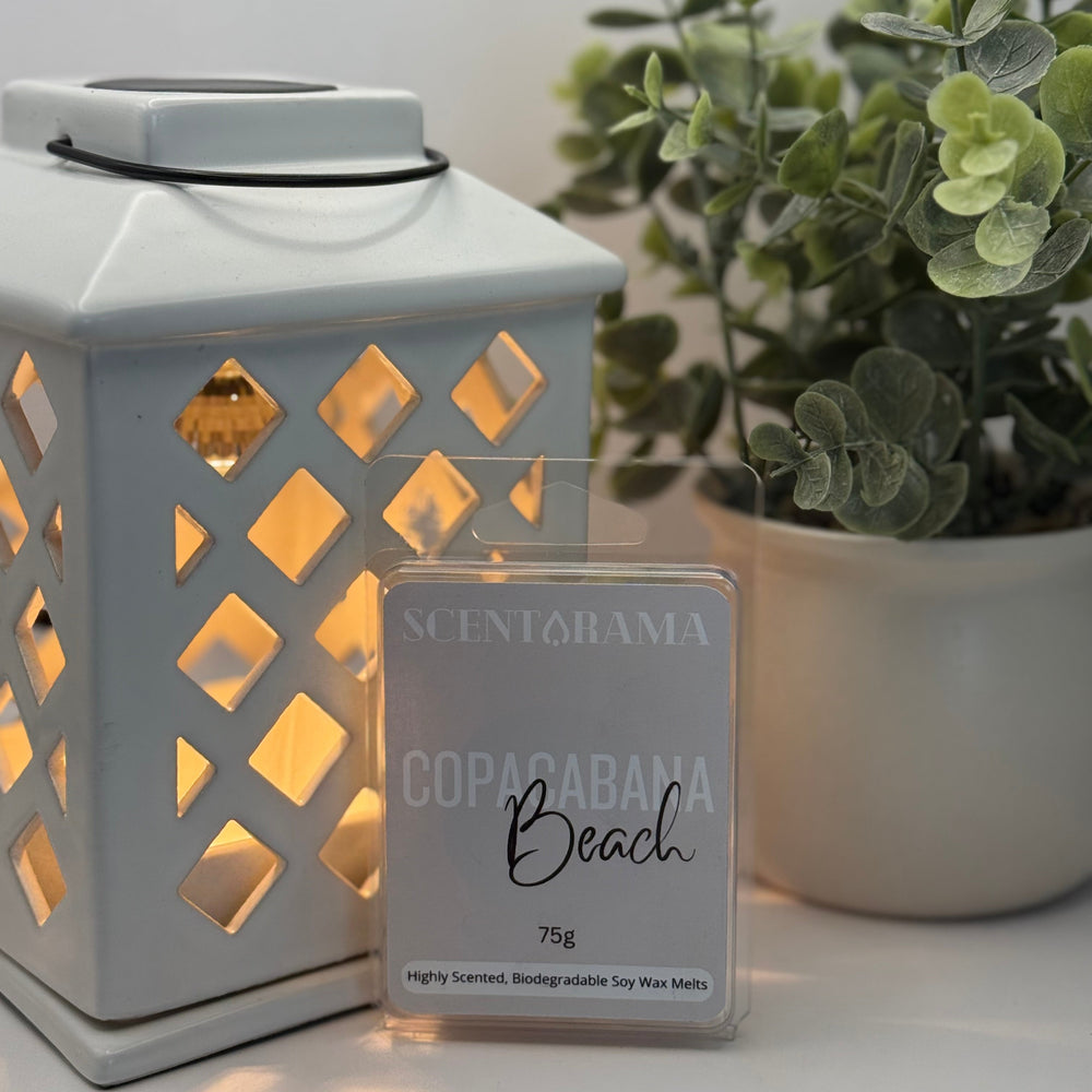 White lantern with a Scentarama wax melt clamshell product labeled 'Copacabana Beach' on a light surface with a plant in the background.