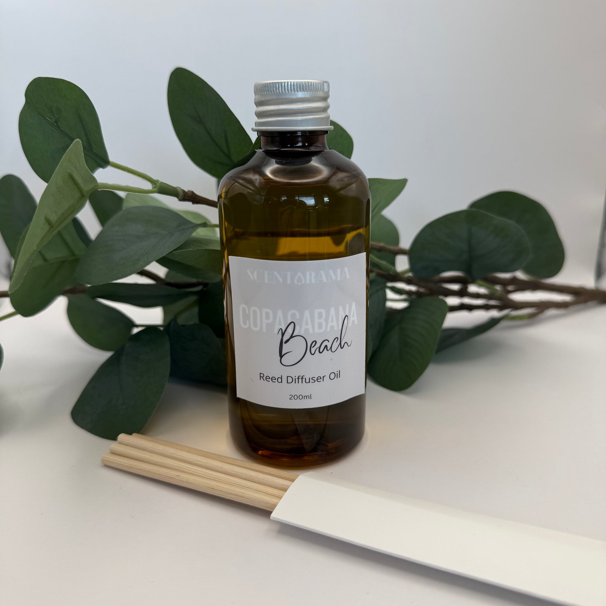 Bottle of Scentarama Copacabana Beach Reed Diffuser Oil with reeds and packaging on a white surface with green leaves in the background