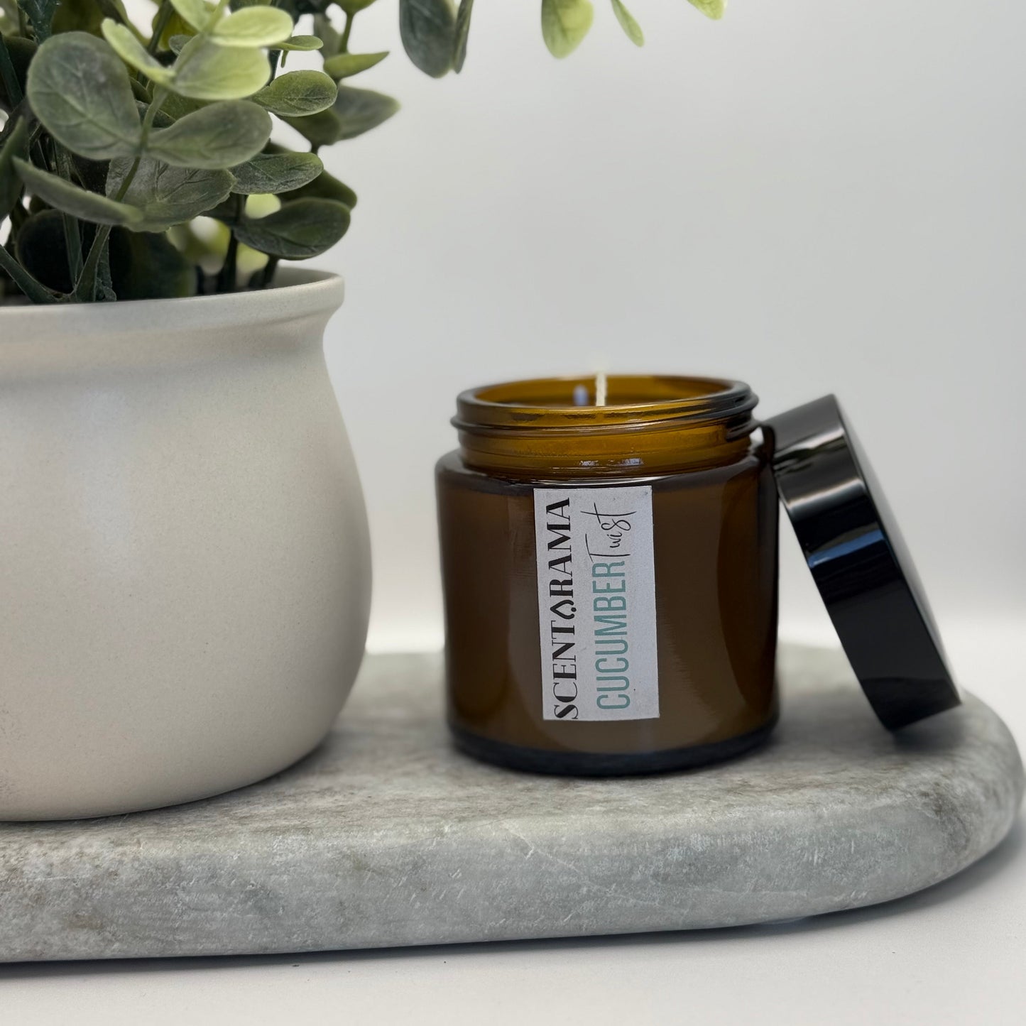 Amber candle with black lid in Cucumber Twist Scent, on a gray tray next to a white pot, featuring a Scentarama label