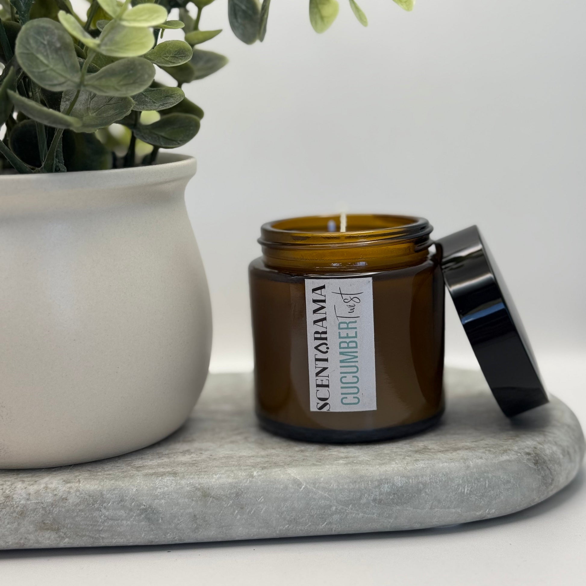 Amber candle with black lid in Cucumber Twist Scent, on a gray tray next to a white pot, featuring a Scentarama label
