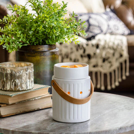 A white ceramic candle warmers Australia fragrance warmer with a leather-look handle and vertical detailing, sitting on a marble surface with a plant and books in the background.