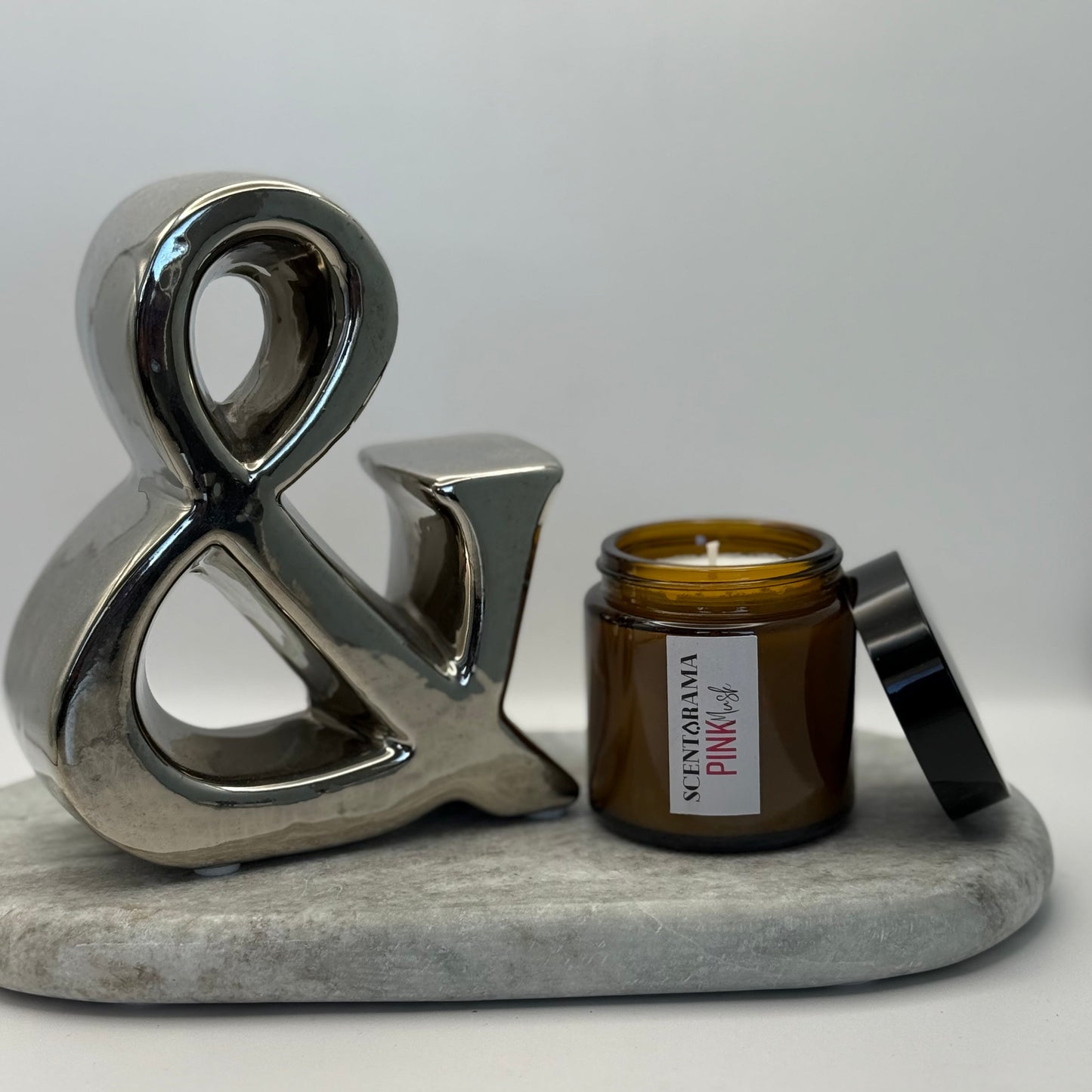 Decorative ampersand and amber candle jar in pink musk scent on a marble base with a white background