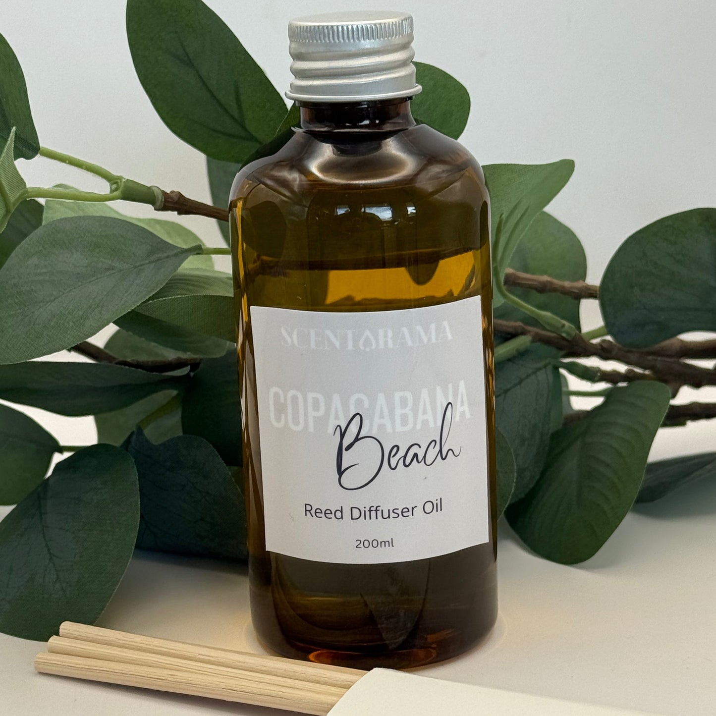 Bottle of Copacabana Beach Reed Diffuser Oil with a label on a white surface with green leaves.