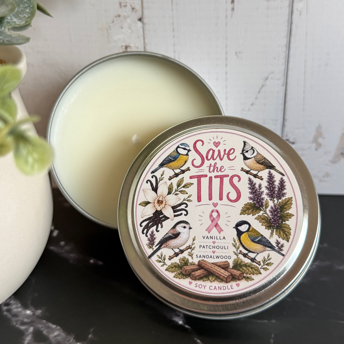 Candle in a tin with decorative label featuring birds and text, on a marble surface.