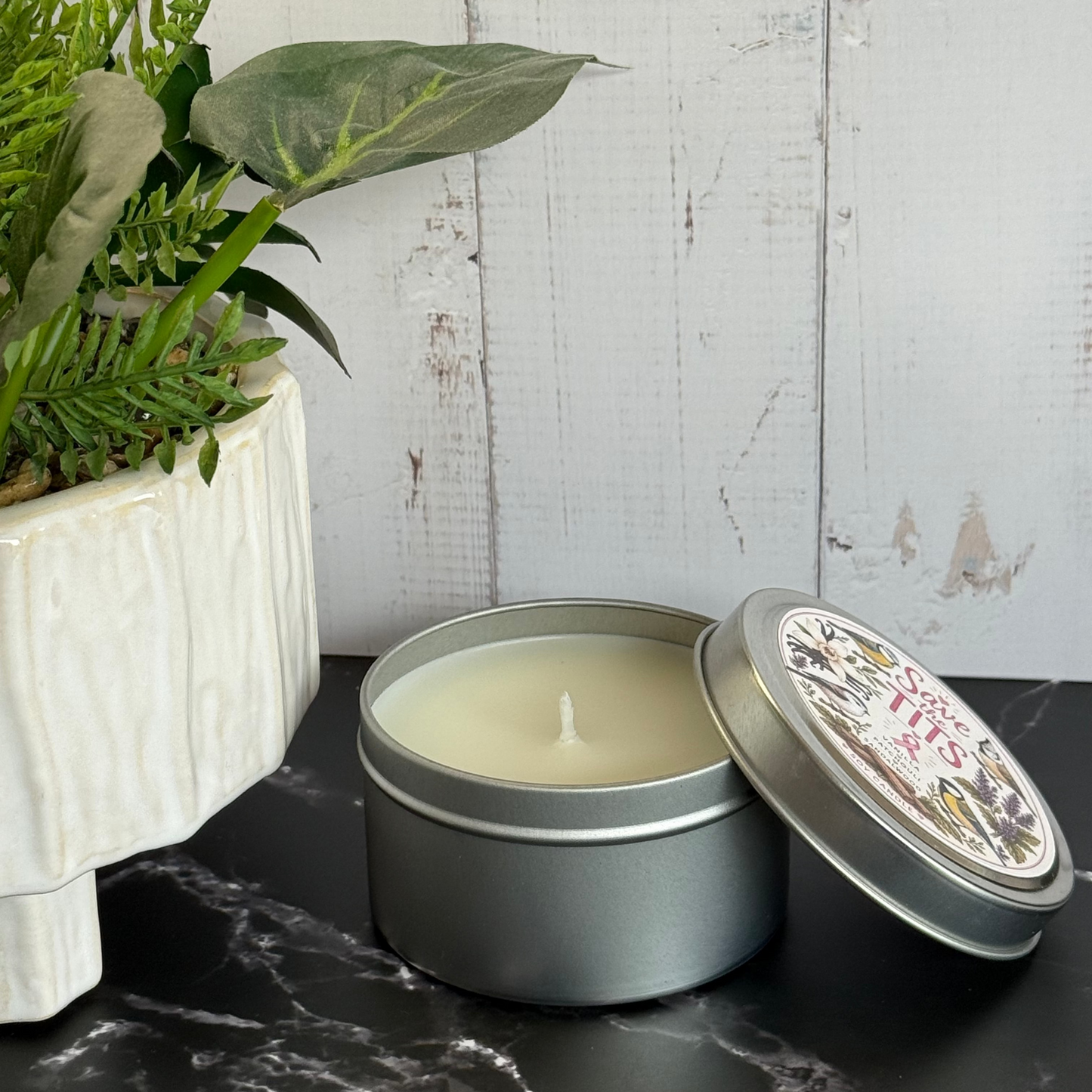 Candle in a tin with a decorative lid on a dark surface next to a potted plant.