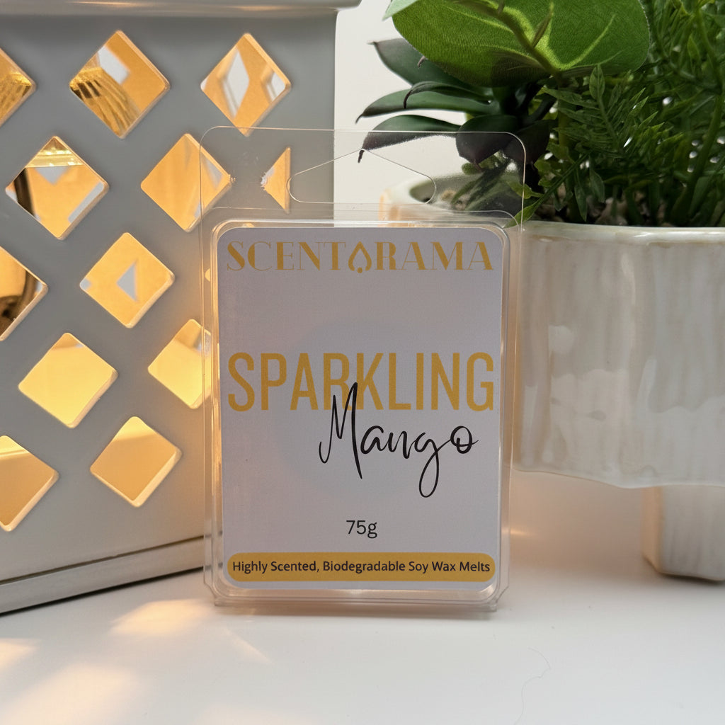 A 75g pack of Scentarama Sparkling Mango soy wax melts in a clear clamshell package, displayed on a white surface next to a lit decorative lantern and a potted plant.