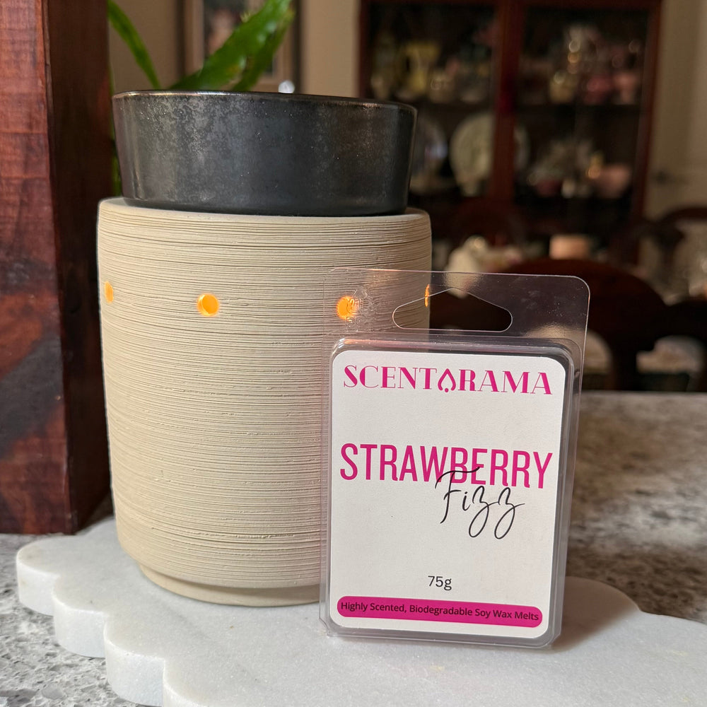A packaged Scentarama Strawberry Fizz wax melt sits in front of a cream-colored ceramic wax melt warmer with small light openings, both placed on a white scalloped tray on a speckled countertop with a blurred indoor background.