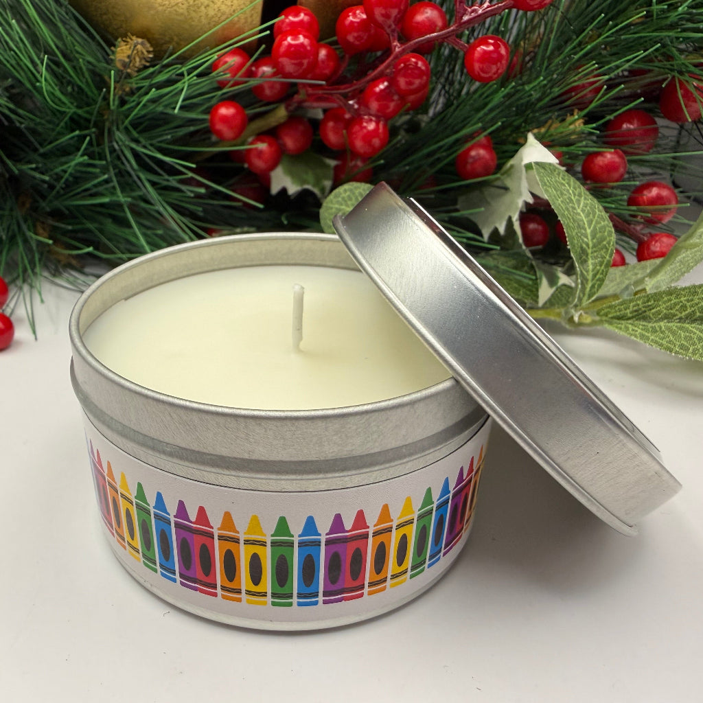 Candle in a tin with colorful crayon design on a white surface with Christmas decorations in the background.
