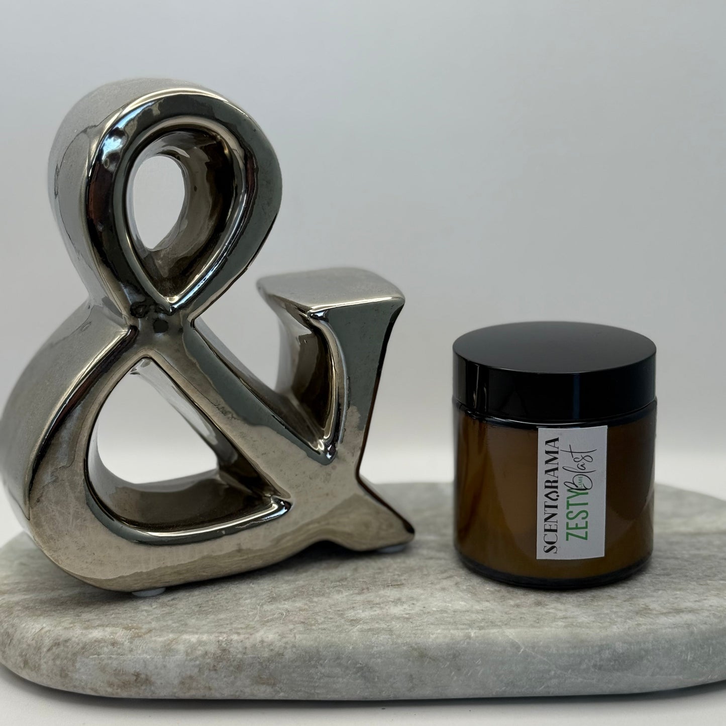 Decorative metal ampersand next to a small amber candle jar in zesty lime blast fragrance featuring a scentarama label sat upon a marble surface with a white background