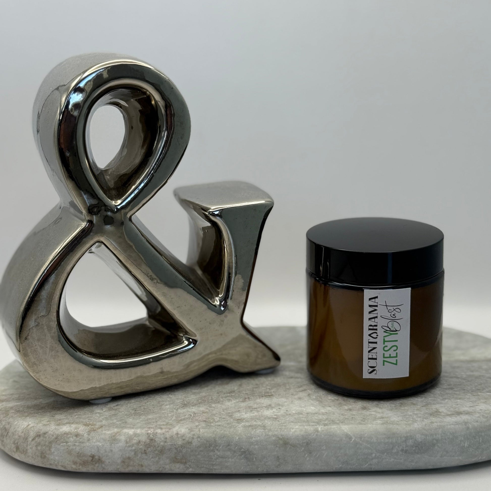 Decorative metal ampersand next to a small amber candle jar in zesty lime blast fragrance featuring a scentarama label sat upon a marble surface with a white background